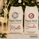 Dos sacos de regalos personalizados con nombres Marta y Sandra y diseños de Papá Noel junto a un árbol de Navidad decorado