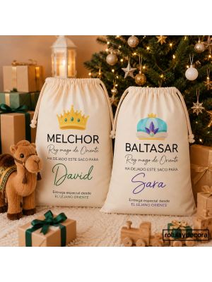 Sacos de regalos personalizados de los Reyes Magos Melchor y Baltasar, de algodón con los nombres David y Sara, junto a un árbol de Navidad decorado, regalos y un camello de peluche.