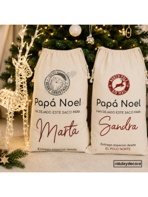 Dos sacos de regalos personalizados con nombres Marta y Sandra y diseños de Papá Noel junto a un árbol de Navidad decorado