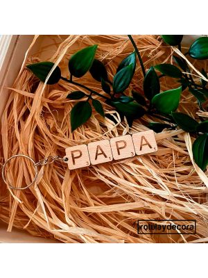Llavero de madera de haya, grabado al láser con letras de Scrabble, formando la palabra Papá,  con anilla y cadena en color acero o plata. Fondo sobre paja y hojas verdes.